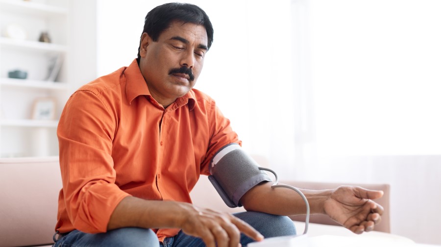 A middle aged Indian man wearing a casual outfit sitting on couch, checking his blood pressure with modern electronic tonometer